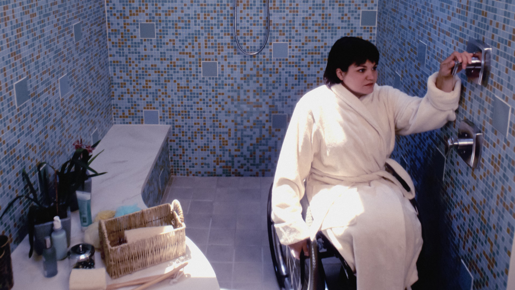 Eine Rollstuhlfahrerin in einer Dusche. Die Dusche ist ebenerdig, sodass die Person problemlos mit dem Rollstuhl in die Dusche fahren kann.