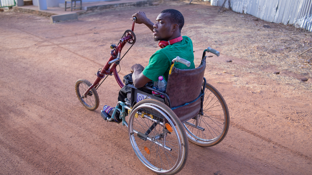 Ein Mann in einem Rollstuhl auf einer unbefestigten Straße. Der Rollstuhl hat nur drei Räder. Das Vorderrad ist mit einem Lenker verbunden. Die beiden Enden der Lenkstangen sind Kurbeln - ähnlich zu Fahrradpedalen - nur dass sie mit den Armen bedient werden können.